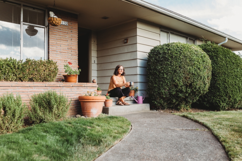 Woman sitting on stoop in front of her house surrounded by green hedges and grass.