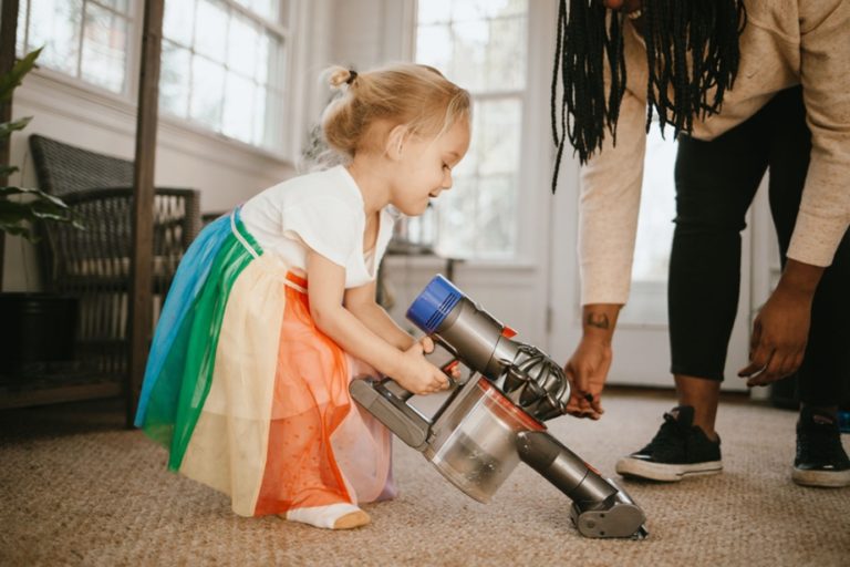 Woman helping toddler girl use a vacuum cleaner.