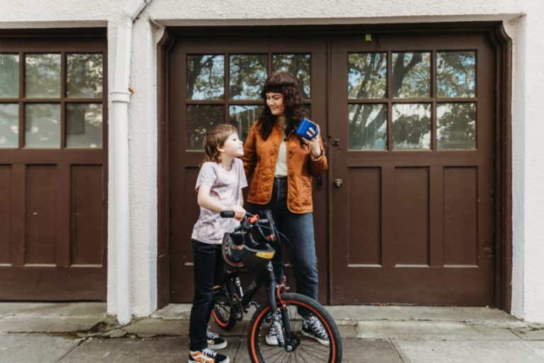 Mom stands with her son while he rides a bike in front of a row of garages.