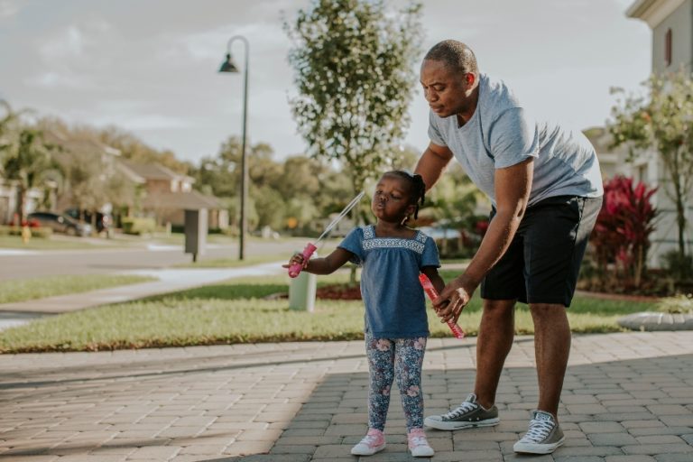 Man helps daughter while she blows bubbles on the sidewalk in front of their home.