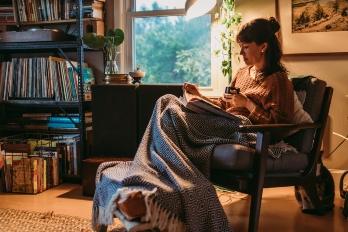 Woman reads a book in a dimly lit living room while snuggled on a chair under a warm blanket.