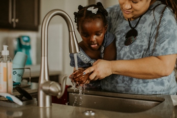 Woman helps her child wash her hands in the kitchen sink.