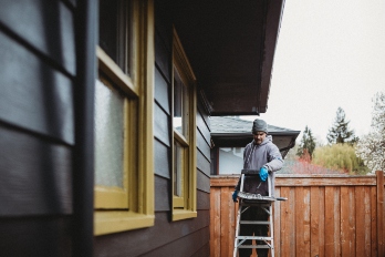 Man stands on a ladder while painting the siding on his house.