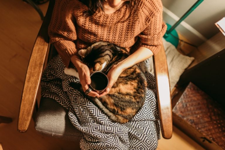 A woman sits under a blanket holding a cup of tea with a cat curled up in her lap.